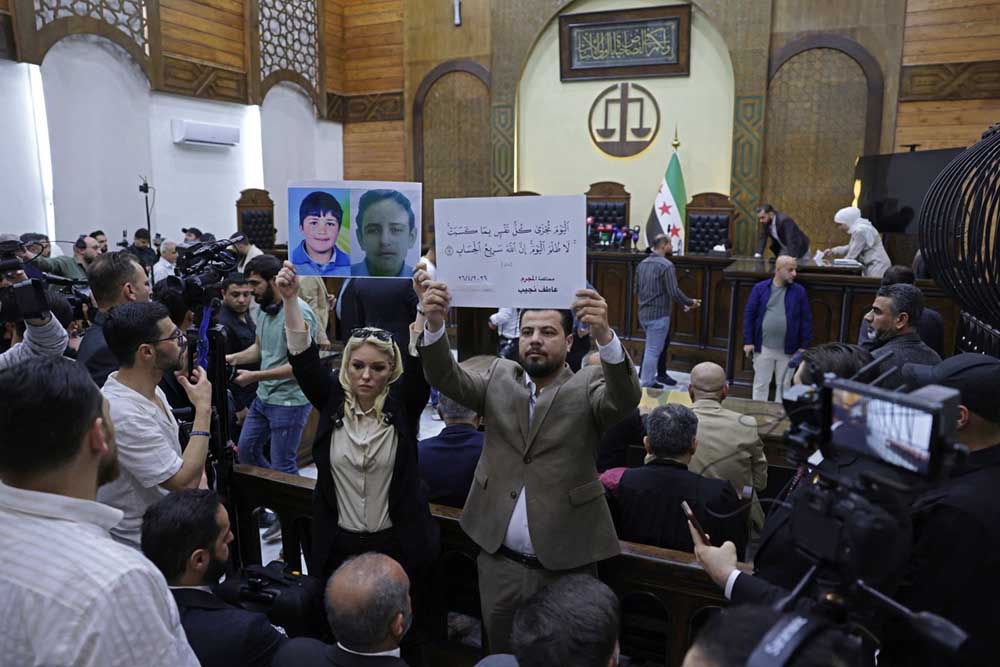  People hold placards at the Palace of Justice ahead of the first trial session, in Damascus on April 26, 2026. (Credit: AFP)