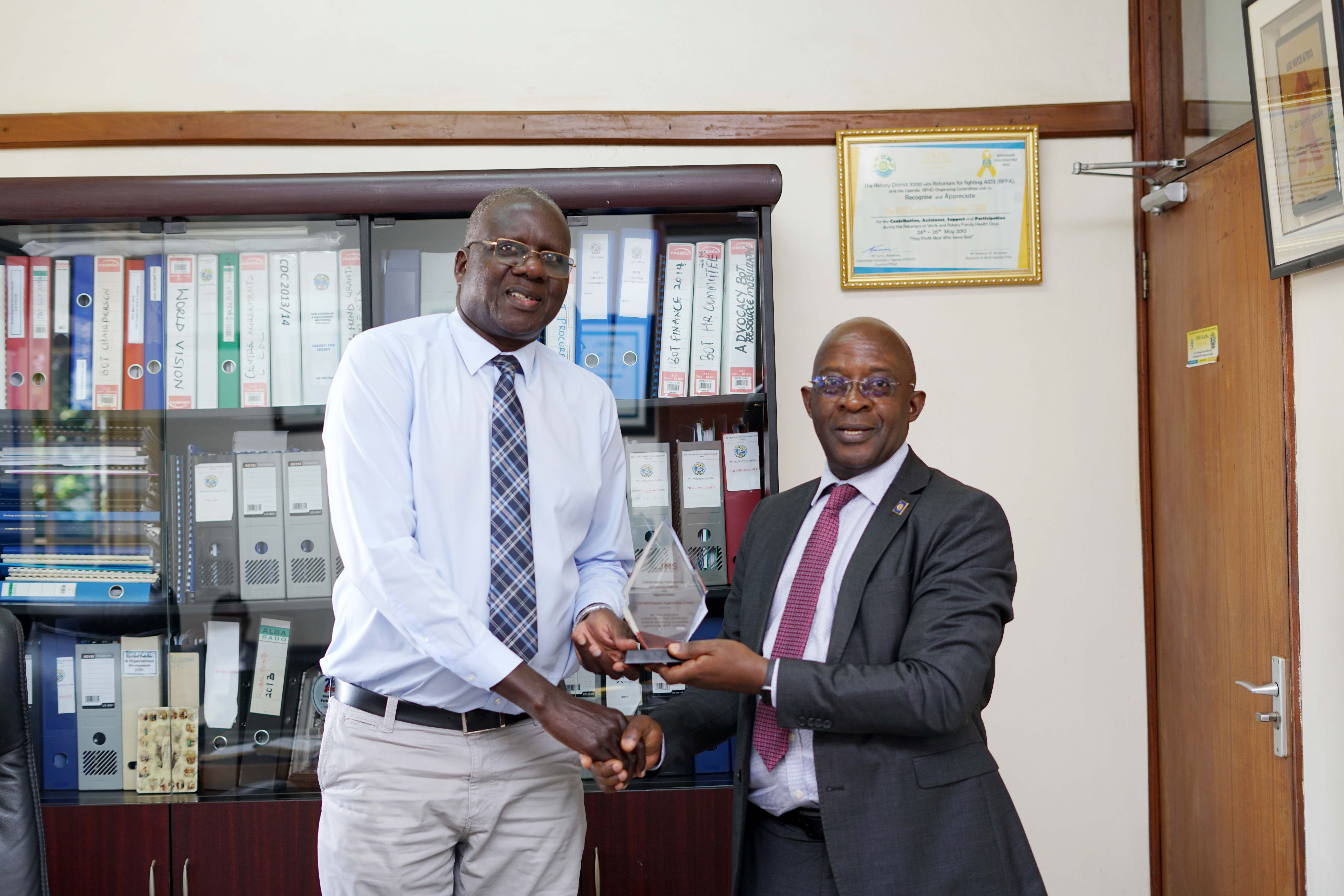 Bildard Baguma (right), the Executive Director of Joint Medical Store (JMS) handing over award to Etukoit Bernard Michael (left), the Executive Director The AIDS Support Organisation (TASO) celebrating partnership honoring impact appreciates during Joint Medical Store meeting The AIDS Support Organistion at The AIDS Support Organisation Mulago on September 4, 2025.