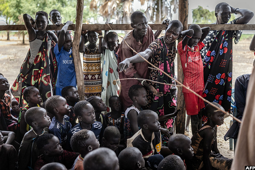 A girl from the Dinka community sits on a tree branch where people have hung their belongings as others gather under its shade at an informal site where displaced people have assembled without assistance or shelter at Yolakot informal camp near Mingkaman, on February 14, 2026