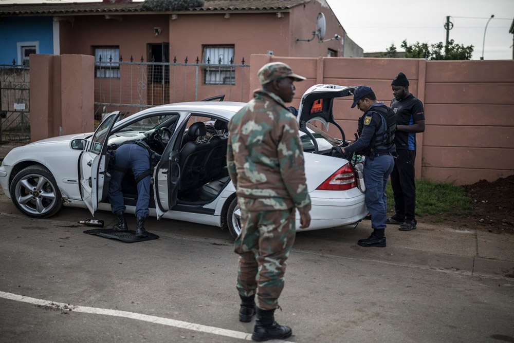 South African Police Service officers conduct a search on the car of a man suspected of harbouring looted items as an officer of the South Africa National Defence Forces (SANDF) stands guard in the Pellsrus township in Jeffreys Bay, on July 18, 2026. (Credit: AFP)
