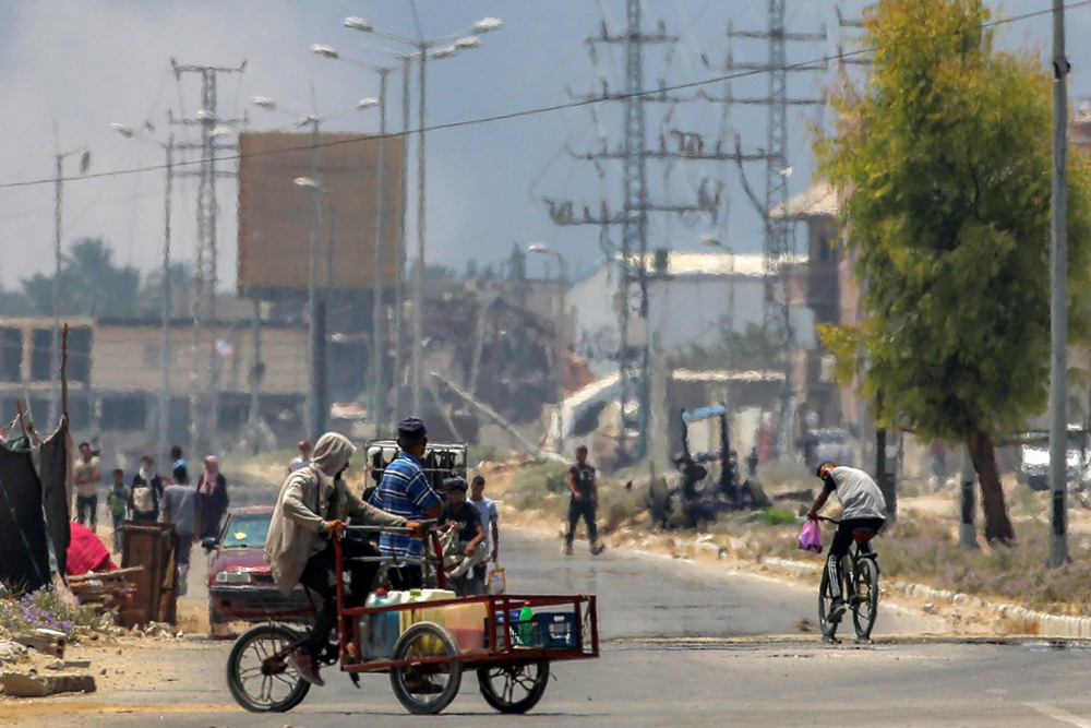 A man on a bicycle speaks with another on a pedal tricycle cart carrying jerrycans as they move along Salaheddin road in Deir el-Balah in the central Gaza Strip on July 21, 2025 while behind smoke billows from Israeli bombardment. 