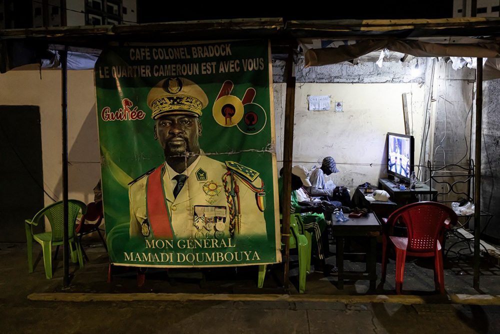 Residents watch television next to a poster depicting Guinea President Mamady Doumbouya who on September 21, 2021, overthrew the elected civilian president, Alpha Condé, who had been in power since 2010. (AFP Photo)