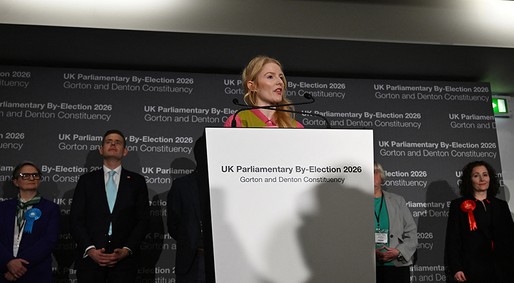 Reform UK candidate Matt Goodwin (2L) and Labour party candidate Angeliki Stogia (R) listen as Green Party candidate Hannah Spencer gives her acceptance speech after winning the Gorton and Denton Parliamentary by-election, at Manchester Central Convention Complex in Manchester, northern England on February 27, 2026. (Credit: AFP)