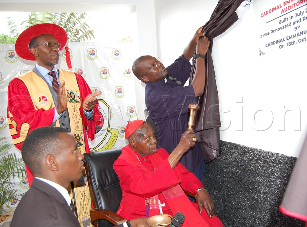 Emmanuel Cardinal Dr Wamala (wearing a skull-cap) sprinkles holy water on the wall of the building that houses the newly-renovated UMU auditorium that was named after him. Looking on is the Vice-Chancellor, Prof. Patrick Kyamanywa (donning academic robes). (Photo by Mathias Mazinga)