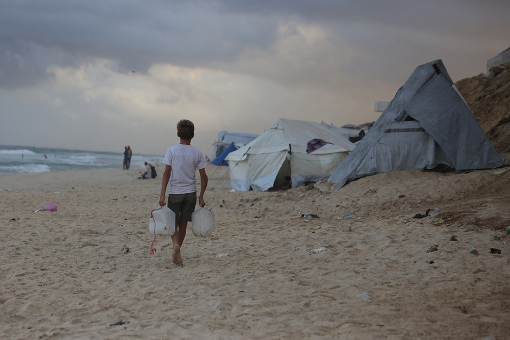 Nizar, 14, a displaced Palestinian boy from the Naqla family in Jabalia in the northern Gaza Strip walks back to his temporary family cave by the beach after fetching drinking water from a distribution point outside a camp in Al-Zawayda city, near Deir al-Balah, in central on October 9, 2025. The family of seven, who lost a 17-year-old son in Israeli strikes in northern Gaza, were displaced earlier this year from Jabalia to the Shati refugee camp in Gaza City, which they had to evacuate last month under Israeli military orders to the central Gaza Strip amid a major ground offensive in the area. 