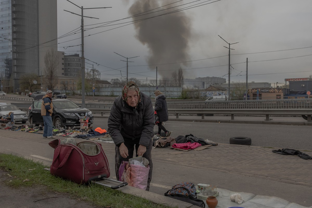 An elderly woman prepares goods at a flea market as smoke rises in the distance following a Russian missile attack, in Kyiv on April 6, 2025, amid the Russian invasion of Ukraine.
