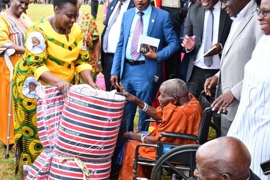 Vice President Maj. (Rtd) Jessica Alupo (C) handing over a mattress and a suitcase gift to 112-year-old Peruth Kyazike (seated right) during celebrations to mark the International Day of Older Persons at St. John Wakitaka SS playground.